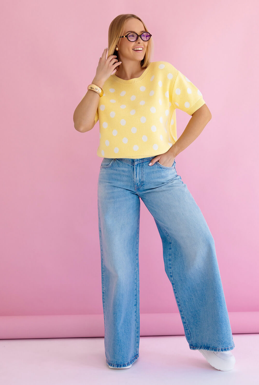 Dame in gelbem Punkte Strickpullover mit kurzen Ärmel mit high-waisted Jeans vor pinkem Hintergrund.